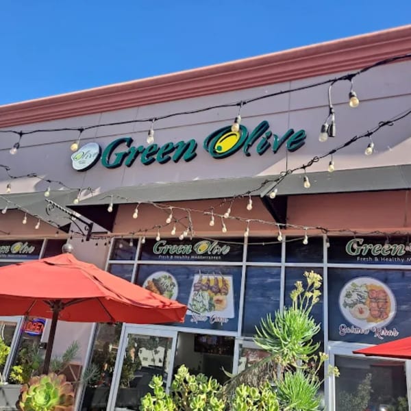 The Green Olive restaurant exterior with a large sign, string lights, red umbrellas, and menu posters.