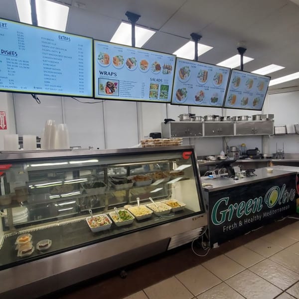 A Green Olive restaurant interior with a food display case, menu boards, and kitchen area.