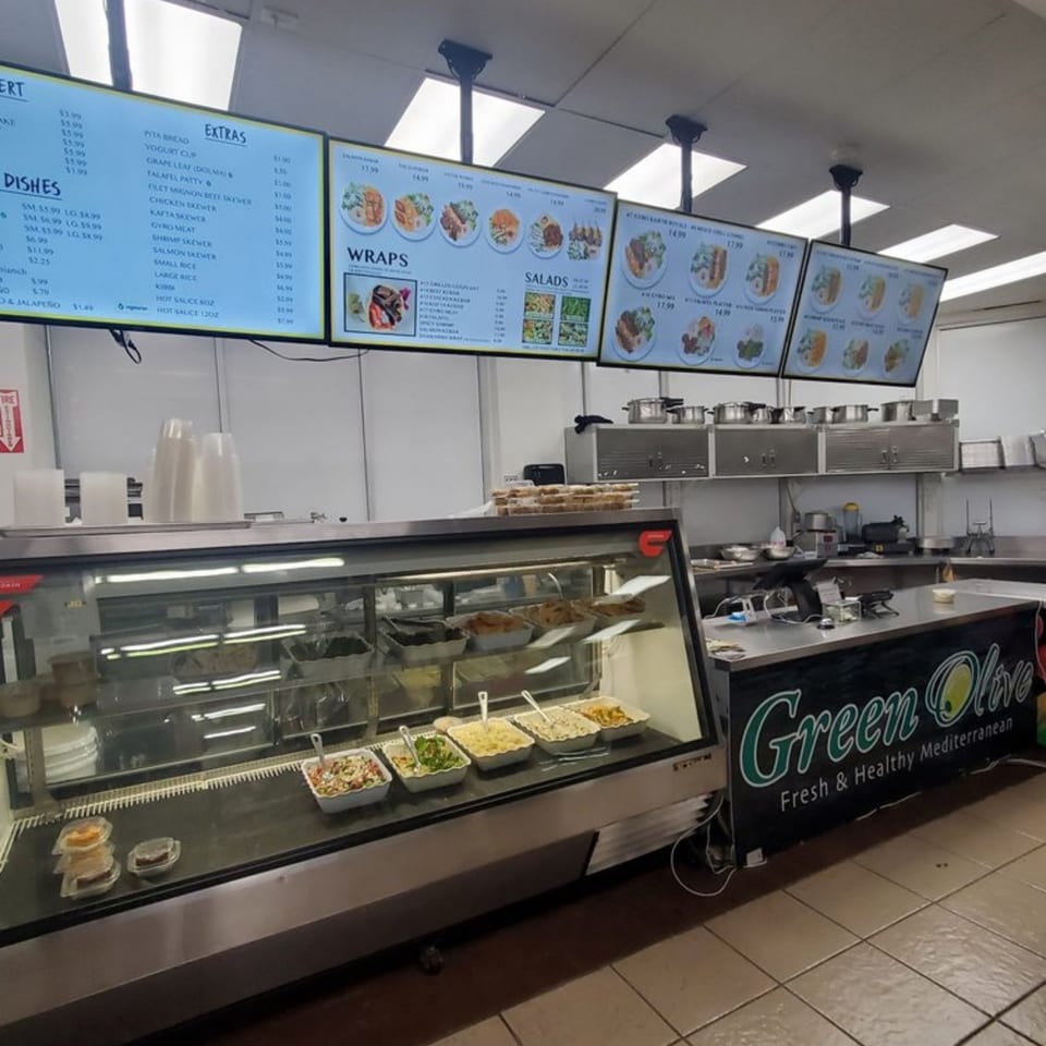 A Green Olive restaurant interior with a food display case, menu boards, and kitchen area.