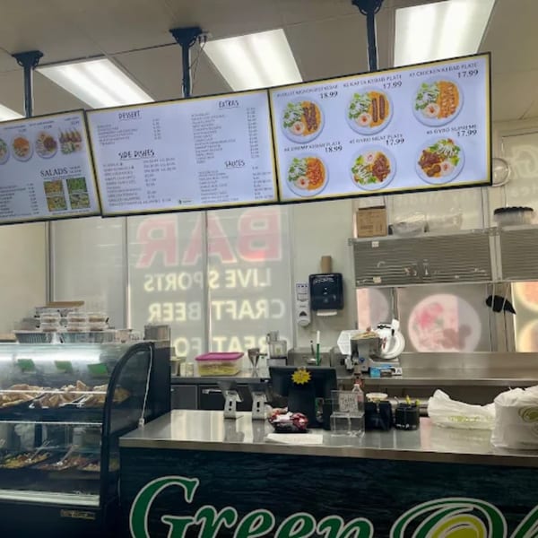 A restaurant counter with a display case of food, digital menus above, and a "Green" logo.
