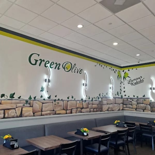 Interior of Green Olive restaurant with "Green Olive" and "Famous Kabobs" signs, stone wall decor, and empty tables.