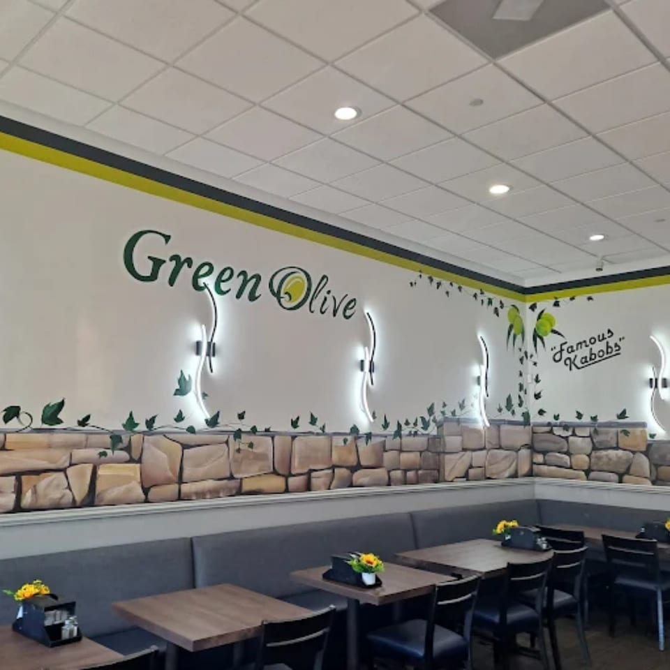 Interior of Green Olive restaurant with "Green Olive" and "Famous Kabobs" signs, stone wall decor, and empty tables.