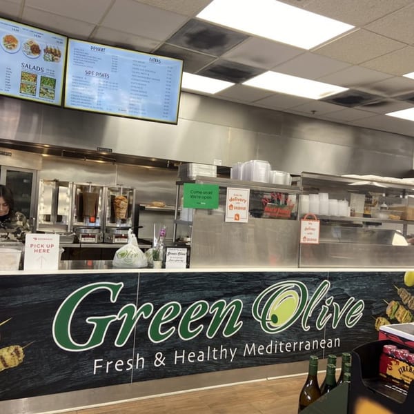 A Green Olive restaurant counter with a menu board, shawarma machines, and a "Pick Up Here" sign.